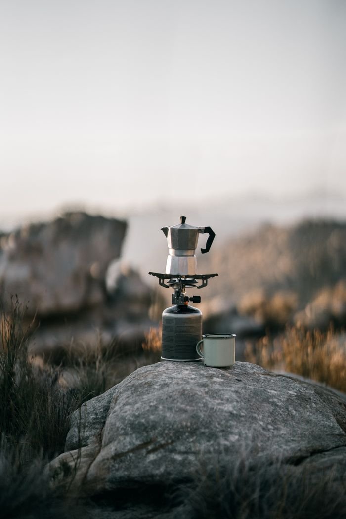 A coffee pot brewing outdoors on a rocky mountain landscape during an early morning hike.